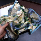 A hand lifting the camellia square, blue border with a fine navy line along the edge