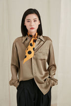 Golden dotted silk scarf knotted long over a khaki satin shirt against a cream curtain