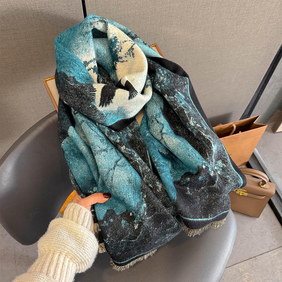 Hand holding the shawl gathered on a grey chair showing the teal and near black marbling and the fringe