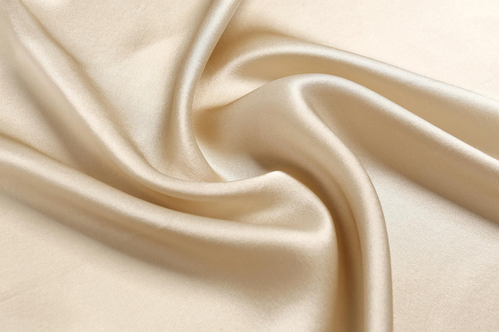 Macro of champagne beige charmeuse swirling into soft folds with a warm satin sheen