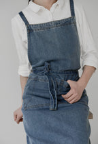 Mid blue denim apron laid flat showing the front pocket and the long shoulder straps