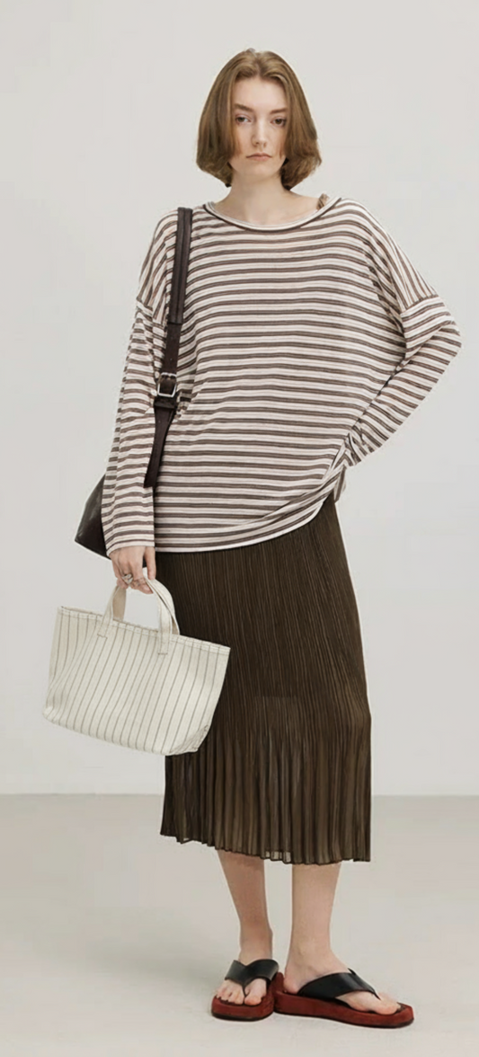 Woman wearing a striped sweater and pleated skirt holding a handbag on a plain background